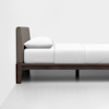 A minimalist bed with a dark wooden frame, white bedding, a single white pillow, and a gray upholstered headboard, set against a plain white wall and floor.