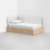 A modern daybed with a light wood base and white upholstered backrest is placed in the corner of a minimalist room. The bed has a white mattress and a single white pillow.