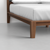 A close-up of a wooden bed frame corner with tapered legs on a smooth, light gray floor. The bed is made with white sheets and pillows, positioned against a white wall.