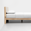 A minimalist wooden bed frame with a light padded headboard, dressed in white bedding and a white pillow, positioned against a plain white wall on a light-colored floor.