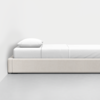 A minimalist single bed with a white frame, white bedding, and a single white pillow, positioned against a plain white wall on a light grey floor.