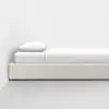 A minimalist single bed with a white frame, white bedding, and a single white pillow, positioned against a plain white wall on a light grey floor.
