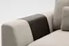 Close-up of a modern beige fabric sofa with a textured, dark brown leather armrest cover draped over the arm. A matching beige cushion is partially visible. The background is plain and light.