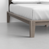 Close-up of the corner of a wooden bed frame with angled legs, supporting a mattress and white bedding in a minimal, modern room with a light gray floor.