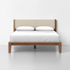 A modern wooden bed frame with a beige upholstered headboard, neatly made with white bedding and two white pillows, set against a plain white wall and floor.