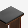 A close-up of a wooden stool with a dark gray cushioned seat, set against a plain white background.