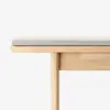 Close-up of a light wood bench with a beige upholstered cushion, shown against a plain white background. Only the top and one leg of the bench are visible.