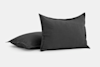 Two dark gray pillows with a soft, linen texture are shown against a plain, light background. One pillow is propped upright, while the other lies flat behind it.