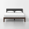A modern bed with a dark wooden frame, white bedding, two white pillows, and a dark gray upholstered headboard against a plain white wall and light gray floor.