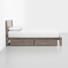 A modern wooden bed with a light gray frame, white bedding, and two large storage drawers underneath, positioned against a plain white wall in a minimalist room.