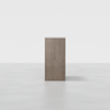 A minimalist wooden cabinet with a light brown finish, viewed from the front, standing alone on a light gray floor against a plain white background.
