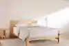 A minimalist bedroom with a light wooden bed frame, white pillows, a light gray comforter, a wooden nightstand with books, and a white floor lamp on a neutral rug against a plain, light-colored wall.