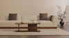 A modern living room with a beige sofa, two dark throw pillows, a wooden coffee table holding a small plant and bowl, and a large potted plant in the corner against a light-colored wall.