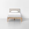 A neatly made single bed with white bedding, a matching pillow, a light beige upholstered headboard, and a simple wooden frame, set against a plain white wall and gray floor.