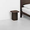 A modern, dark wood nightstand with a flat square top and rounded edges sits next to a matching wooden bed with white bedding in a minimalist room with white walls and floor.