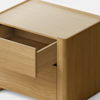 A minimalist wooden nightstand with a single drawer partially open, placed on a light surface against a plain white background.