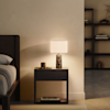 A modern bedroom nightstand with a marble-base lamp, a small black container, and a pair of glasses. The bed is partially visible on the left, and shelves with decor are blurred in the background. Warm lighting creates a cozy atmosphere.