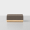 A modern, rectangular ottoman with a brown upholstered cushion and a light wooden base, placed on a light gray floor against a plain white background.