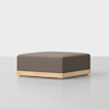 A modern square ottoman with a brown upholstered cushion and a light wooden base, set against a plain white background.