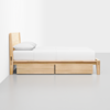 A minimalist wooden bed with a light wood frame, white bedding, and two storage drawers underneath, placed against a white wall and on a light gray floor.