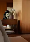 A wooden bedside table holds a black lamp and a potted plant with green and white leaves. Soft natural light highlights the warm wood tones and modern, minimalist bedroom decor.