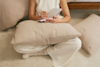 A person in cream-colored clothes sits cross-legged on the floor, writing in a notebook that rests on a beige pillow. Another pillow is nearby atop a light carpet, with part of a bed visible in the background.
