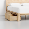 A light wood bed frame with a white mattress and pillows, featuring an open under-bed storage drawer on the side, placed on a smooth, light-colored floor.
