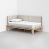 A minimalist daybed with a light wood frame, beige upholstered backrest, and a white mattress with a pillow, placed in the corner of a plain white room.