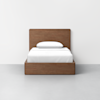A wooden bed frame with a headboard and storage base, shown from the front, with a neatly made white bed and a single white pillow against a plain white wall and light gray floor.