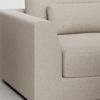 Close-up of a modern beige fabric sofa showing the armrest, seat cushion, backrest, and a rectangular pillow. The upholstery has a subtle textured pattern and the sofa sits on a light floor.