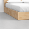 A close-up view of the corner of a light wooden bed frame with white bedding and two pillows, set on a smooth, light grey floor in a minimalistic room.