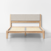 A minimalist wooden bed frame with a light fabric-upholstered headboard, slatted base, and angled legs, set against a plain white wall on a smooth light gray floor.