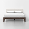 A modern bed with a dark wooden frame, white upholstered headboard, white bedding, and two white pillows, set against a plain white wall and floor.