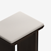 A close-up view of a stool with a dark wooden frame and a light beige, cushioned fabric seat, photographed against a white background.