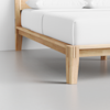 Close-up of a wooden bed frame with a light finish, featuring clean lines and sturdy legs, supporting a mattress with white bedding in a minimal, modern room.