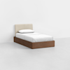 A single bed with a beige upholstered headboard, white bedding, and a wooden base sits against a plain white wall on a light gray floor.