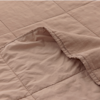 A close-up of a light brown or beige quilt with visible stitching in a grid pattern and one corner gently folded over, showing the soft texture of the fabric.