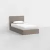 A single bed with a light wood frame, white bedsheet, and white pillow is positioned against a plain white wall on a light gray floor in a minimalist setting.