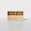 A minimalist wooden cabinet with two open shelves and two lower drawers, placed on a light grey floor against a white background.