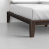 Close-up of the corner of a bed with white bedding and a dark wooden frame, standing on a light gray floor against a white wall.