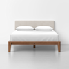 A neatly made bed with white bedding and two pillows, featuring a light upholstered headboard and a simple wooden frame, is positioned against a plain white wall on a light gray floor.