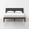 A modern bed with a dark wood frame, black upholstered headboard, white bedding, and two white pillows, set against a plain white wall and floor.
