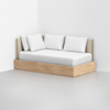 A minimalist daybed with a light wood base, beige upholstered backrest, and four white pillows, placed in a corner of a bright, empty room with light gray flooring.