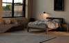 A modern bedroom with a large window, an unmade bed with gray bedding, a sleek floor lamp, a dresser with a water bottle, and a painting on the wall, all in neutral tones.
