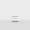 A small, minimalist wooden shelving unit with two shelves, placed in the center of an empty, light-gray room with a plain white wall and a smooth floor.