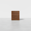A modern, minimalist wooden chest of four drawers is centered on a light gray floor against a plain white background. The chest has a smooth, flat surface with no visible handles.
