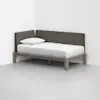 A minimalist daybed with light wood frame, white mattress, white pillow, and brown upholstered backrest is positioned in the corner of a plain white room with a gray floor.