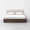A minimalist bed with a light beige upholstered headboard, white bedding, two pillows, and a dark wooden frame, set against a plain white wall and gray floor.