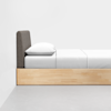 A minimalist bed with a light wooden frame, gray upholstered headboard, white pillow, and white bedding, positioned against a plain white wall on a smooth gray floor.