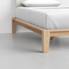 A close-up view of a wooden bed frame with light wood finish and white bedding, showing the corner of the frame and mattress on a light gray floor.